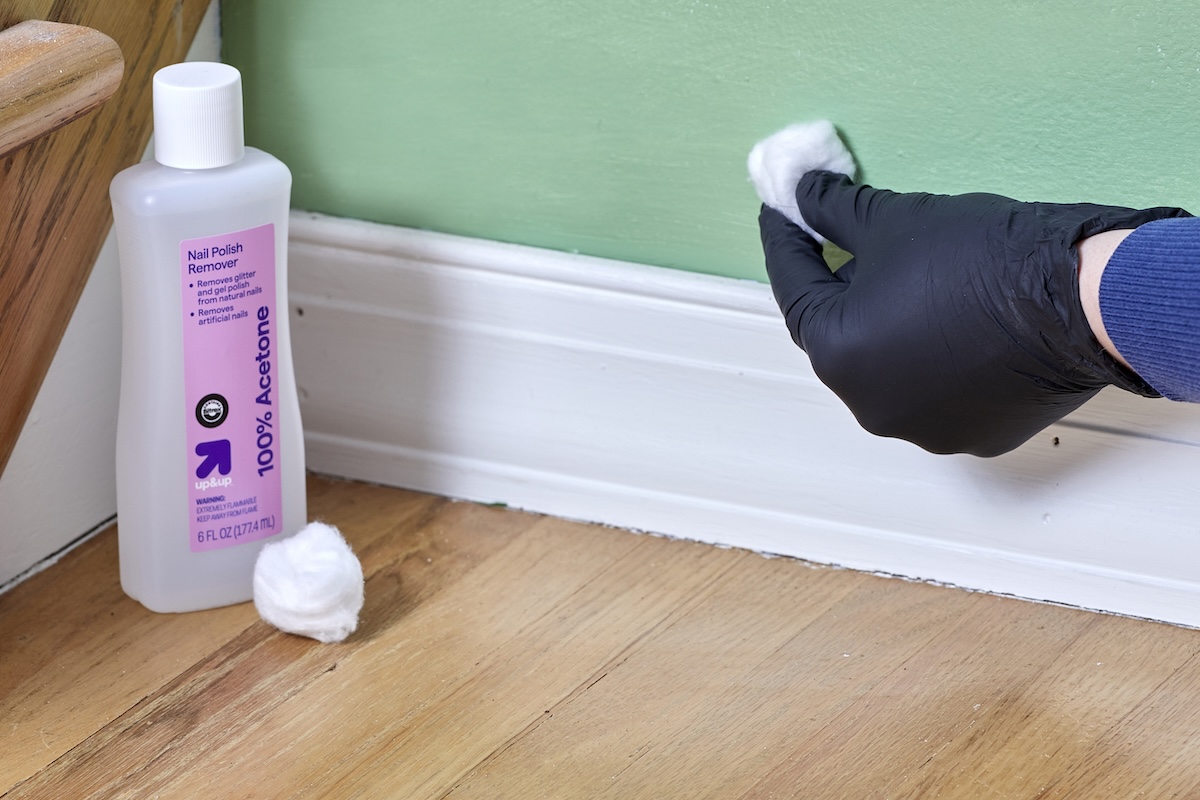 Woman wearing black nitrile gloves rubs a painted wall with a cotton ball dipped in nail polish remover.