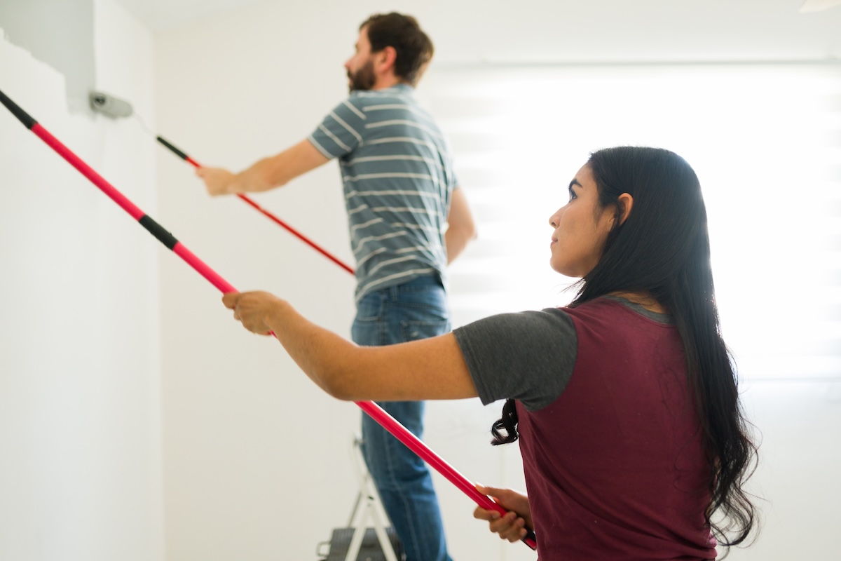 Young couple is painting the walls of their new home gray using paint rollers with extension poles