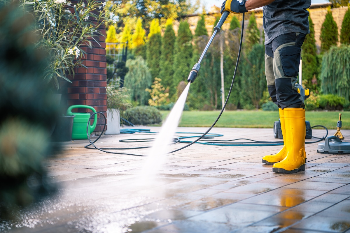 A person wearing yellow boots operates a pressure washer on a patio, creating water spray while surrounded by greenery on a bright day.