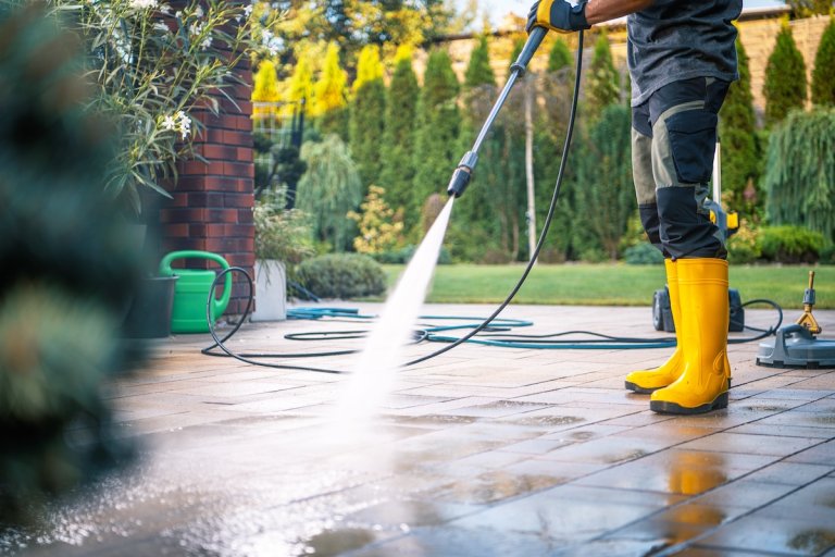 A person wearing yellow boots operates a pressure washer on a patio, creating water spray while surrounded by greenery on a bright day.