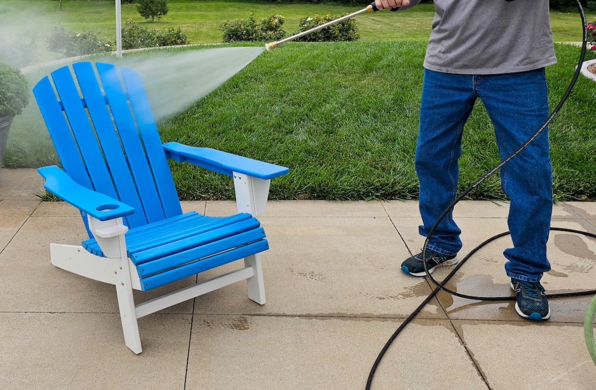 Man wearing jeans uses pressure washer to clean an Adirondack chair painted in blue and white.
