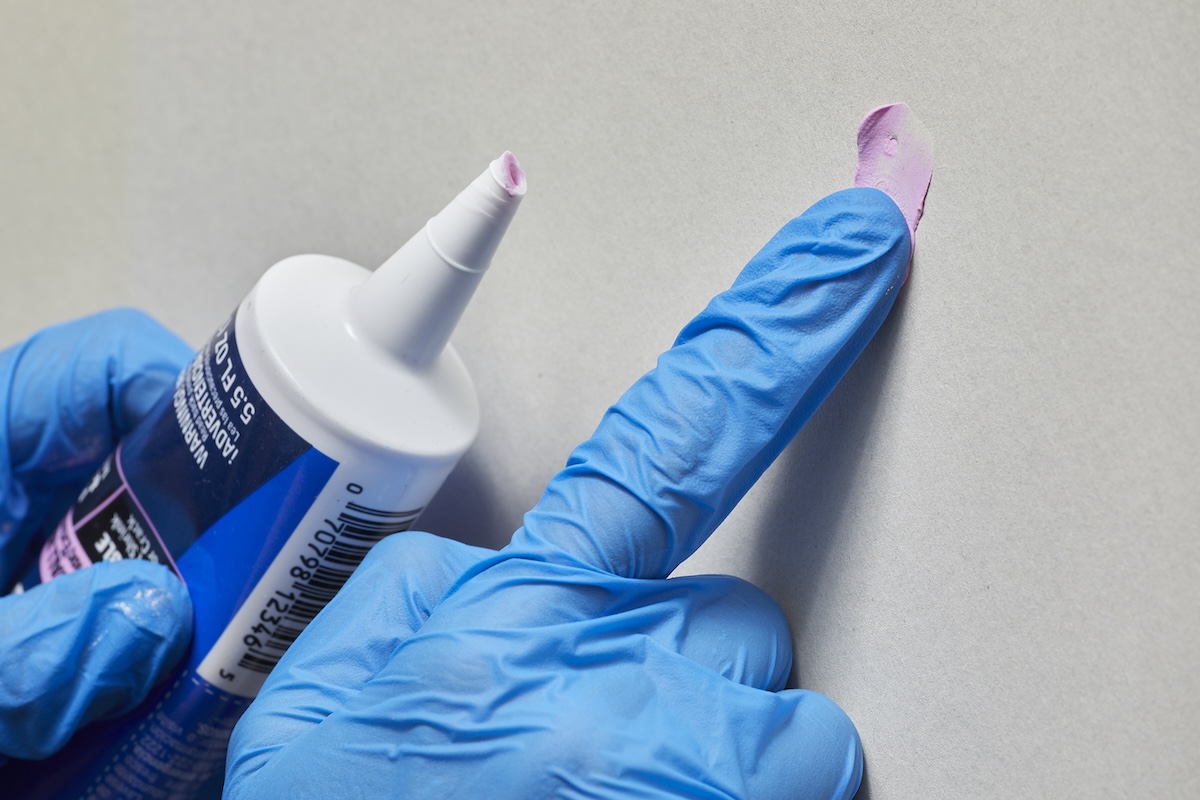 Person wearing blue nitrile gloves puts pink quick-dry spackle on a nail hole with their finger.