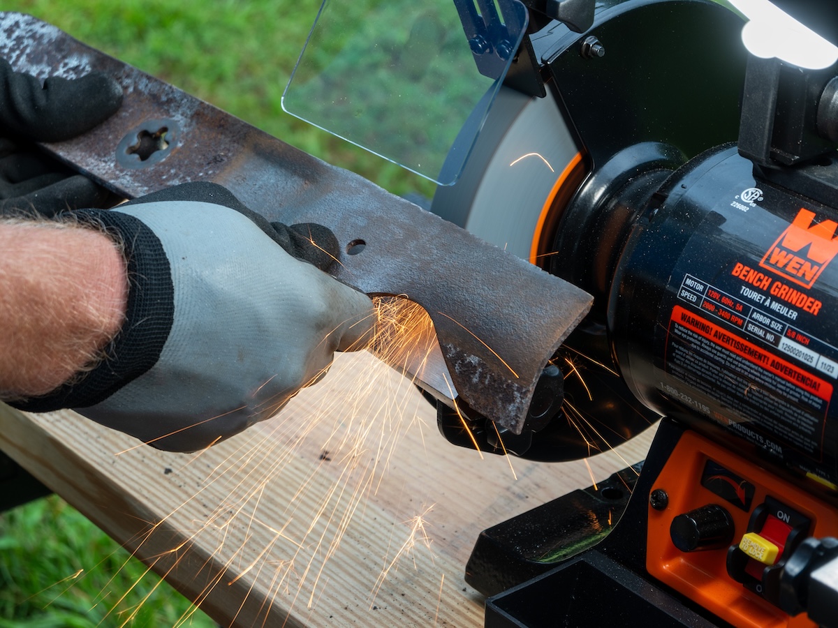 Man wearing work gloves sharpens a lawn mower blade with a blade sharpener on an outdoor table.