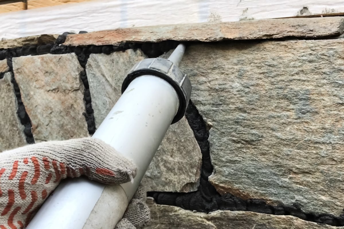 Close up of hand using caulk to fill in gaps between stone on the cladding of the foundation.