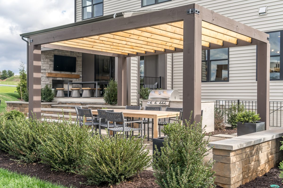 Outdoor patio pergola shade structure, awning and patio roof, dining table, chairs, metal grill surrounded by landscaping.