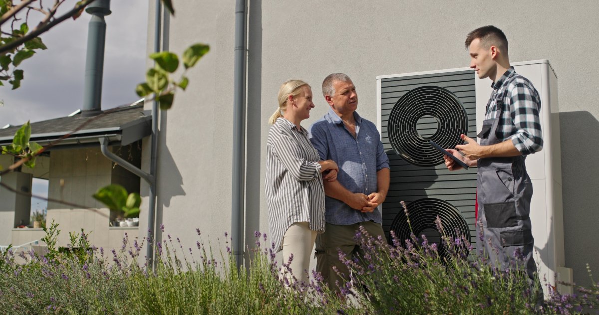A technician uses a tablet to demonstrate heat pump features to a couple outdoors.