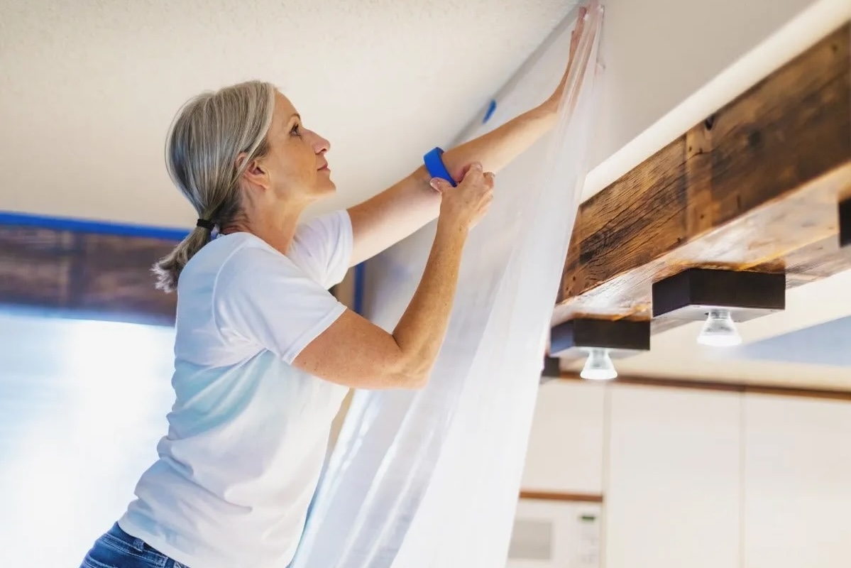 Woman on a ladder hangs plastic sheeting on  a wall near the ceiling, a roll of painter's tape looped around her arm.
