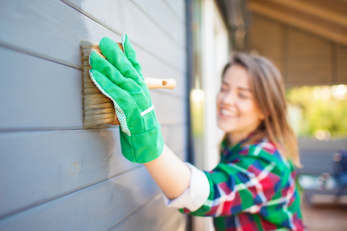 Woman wearing green work gloves paints the exterior of a grey house.