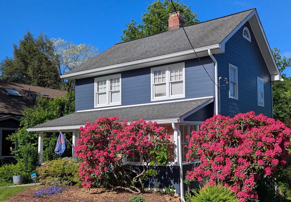 Colonial-style home with dark blue fiber cement siding, with blooming pink rhododendron bushes in the front yard.
