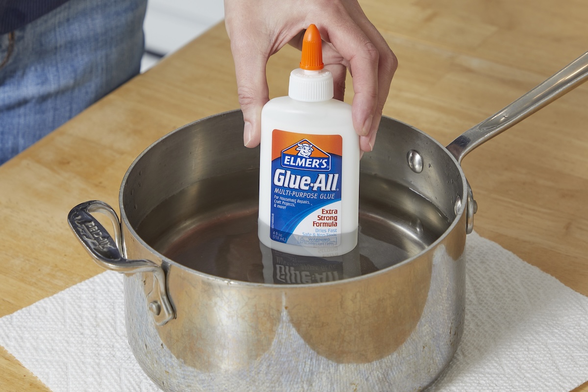 Woman places a bottle of Elmer's Glue-All in a saucepan of water, on top of a paper towel on a countertop.