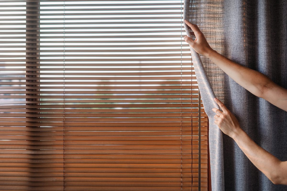 A person opens privacy curtains in front of blinds.