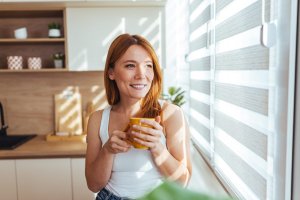 A cheerful woman stands in a bright kitchen, sipping from a yellow mug. Sunlight streams through striped window blinds, creating a warm, relaxed atmosphere.