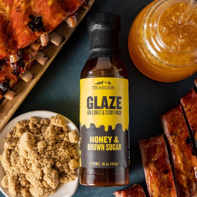 A bottle of Traeger honey and brown sugar glaze next to a rack of ribs, ham, honey, and brown sugar