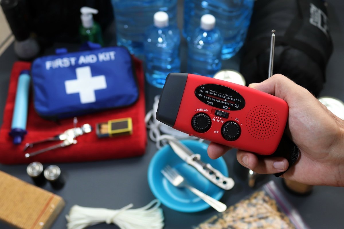 Person holds an emergency radio with other components of an emergency disaster kit in the background (first aid kit, mess kit, etc).