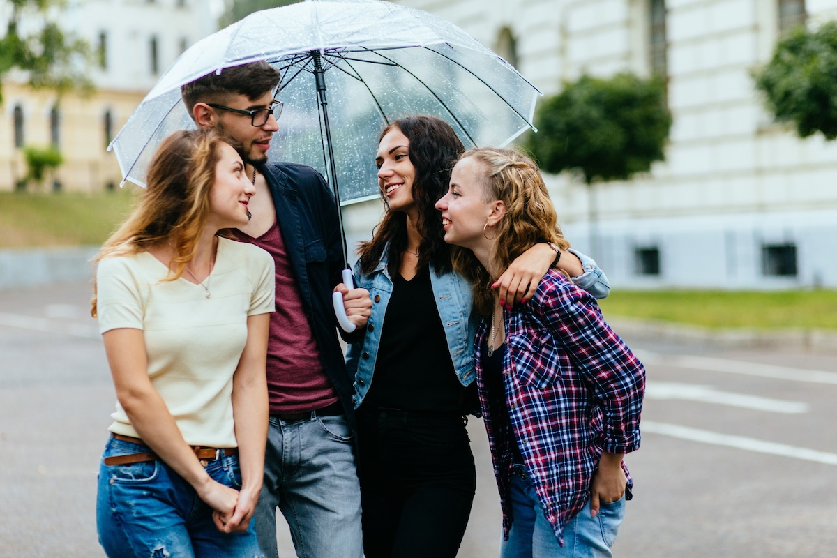 Group of four positive student friends sheltering from rain beneath a big umbrella.