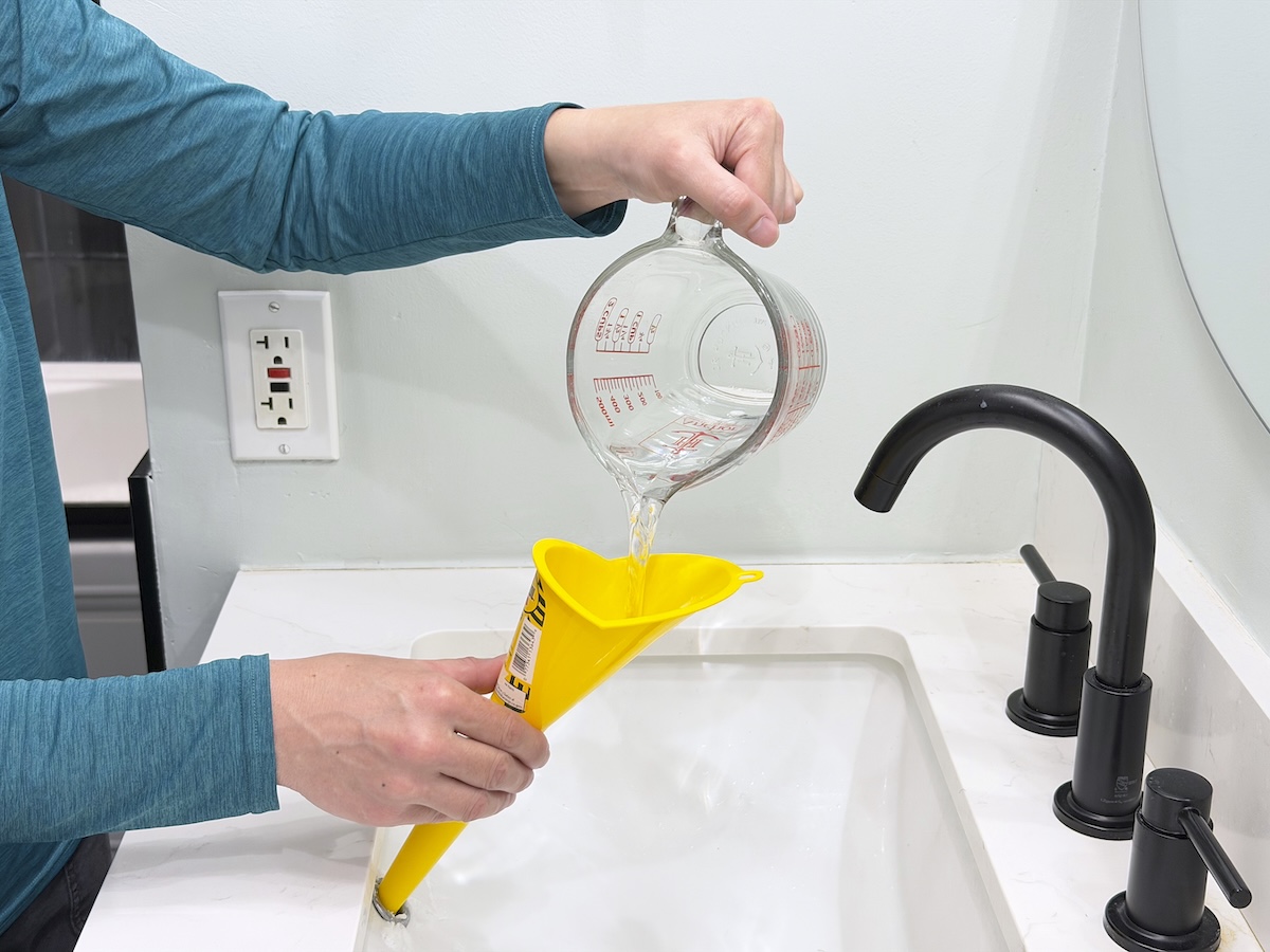 Woman pours vinegar from a measuring cup through a yellow funnel into a hole in the bathroom sink.