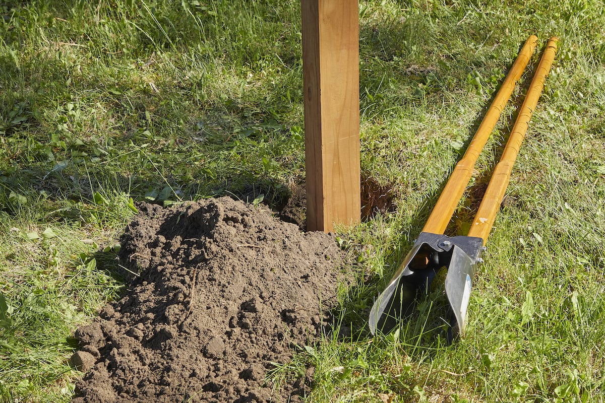 A fence post stands in a freshly dug hole in the ground, soil and the post hole digger next to the post.