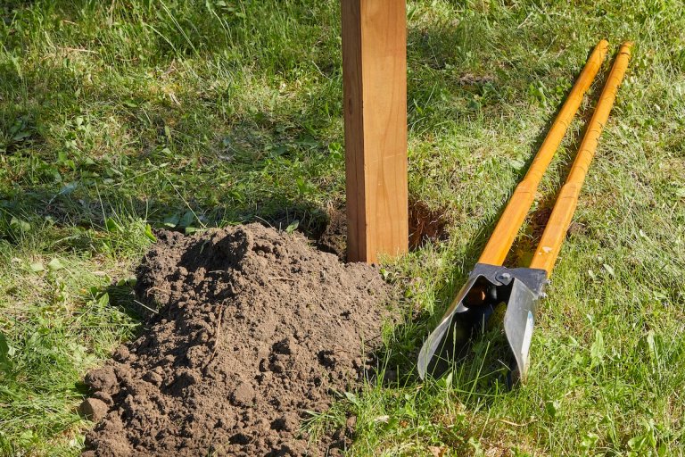 A fence post stands in a freshly dug hole in the ground, soil and the post hole digger next to the post.