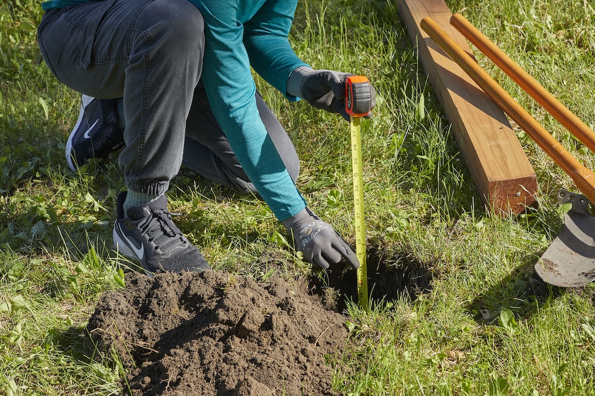 Woman uses a tape measure to measure the depth of a hole in the ground, a fence post and manual post hole digger laying nearby.
