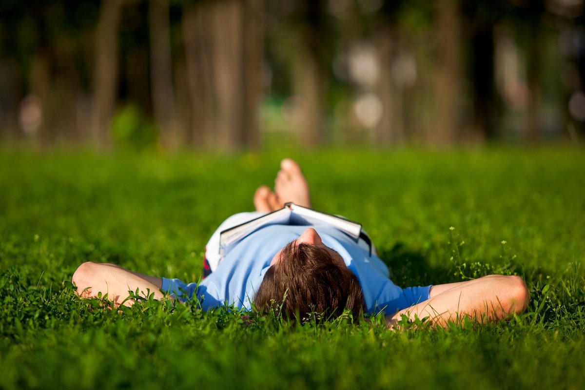 Young man in casual clothing lying on green grass and reading book in park on summer day.