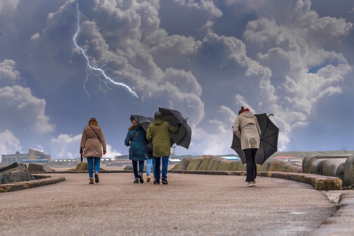 A group of people walking and trying to keep umbrellas in a windy day and lightning in the sky.