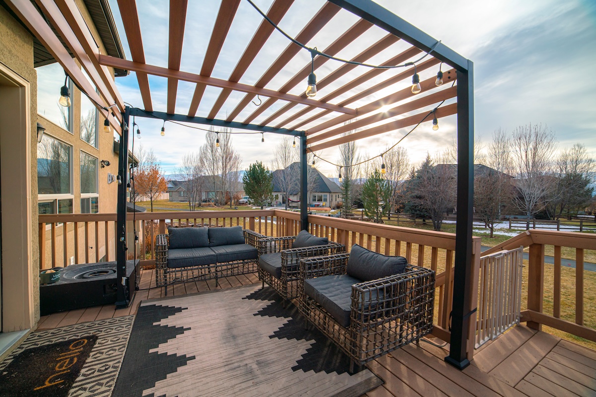 Wooden pergola over an exterior patio with garden furniture and a banister overlooking a large backyard.