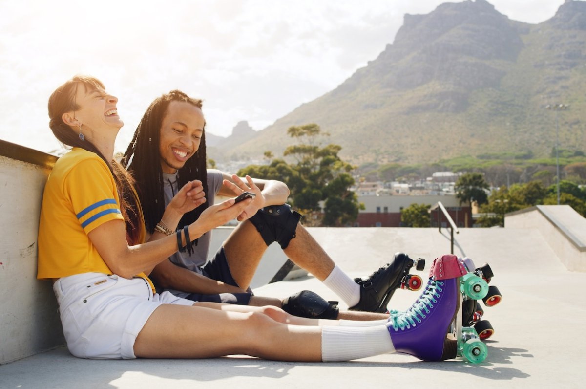Two young people wearing roller skates sit on a concrete walkway looking at a mobile phone and smiling.