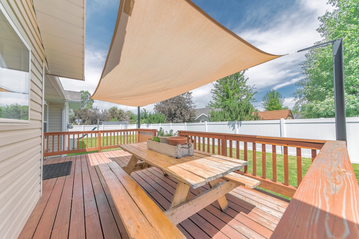 Deck of a house with wooden chairs and table under a shade sail.