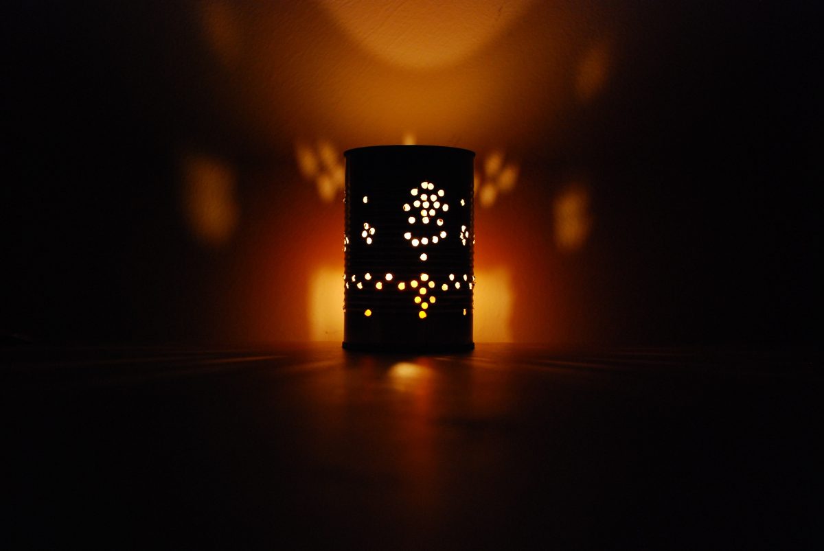 A handmade lantern created from a recycled tin can, lit by a candle inside, casting artistic shadows on the wall and table in a dimly lit room.