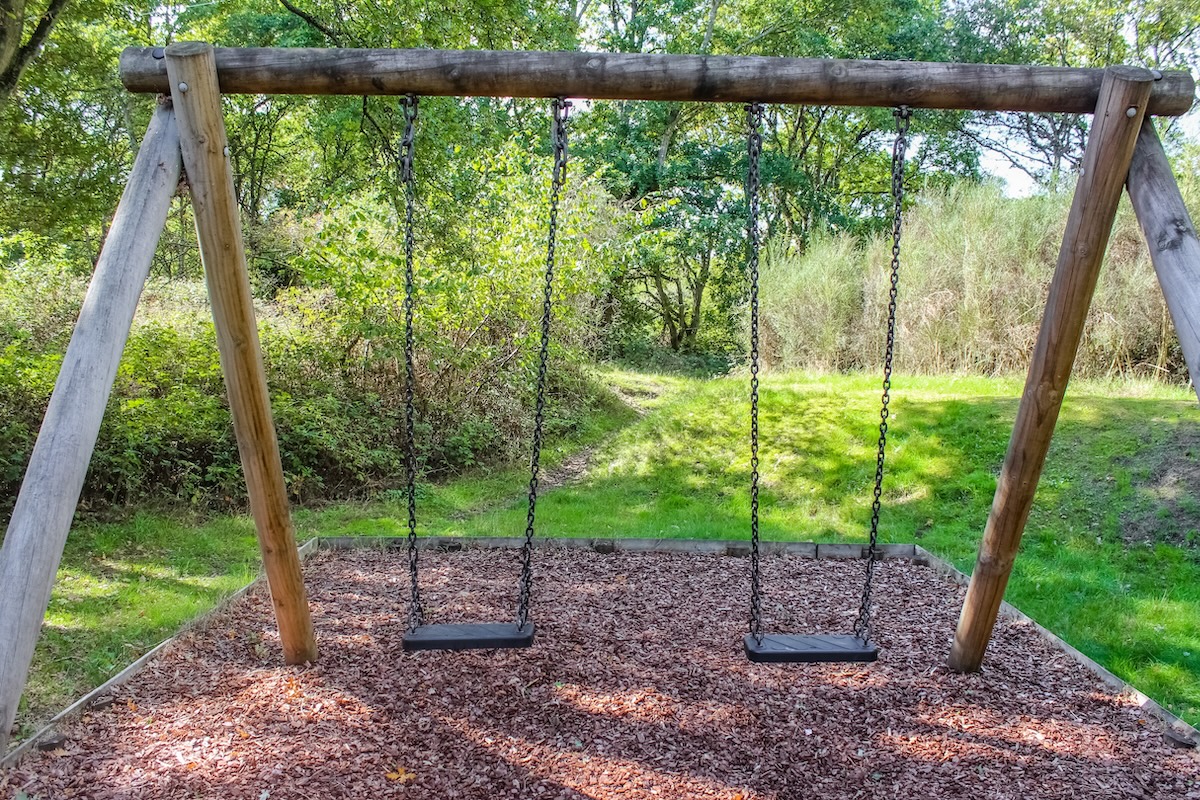 A wooden swing set with two black seats hangs from chains on a sturdy timber frame. It's set in a lush green park over a brown woodchip safety surface, surrounded by dense trees and bright sunlight.