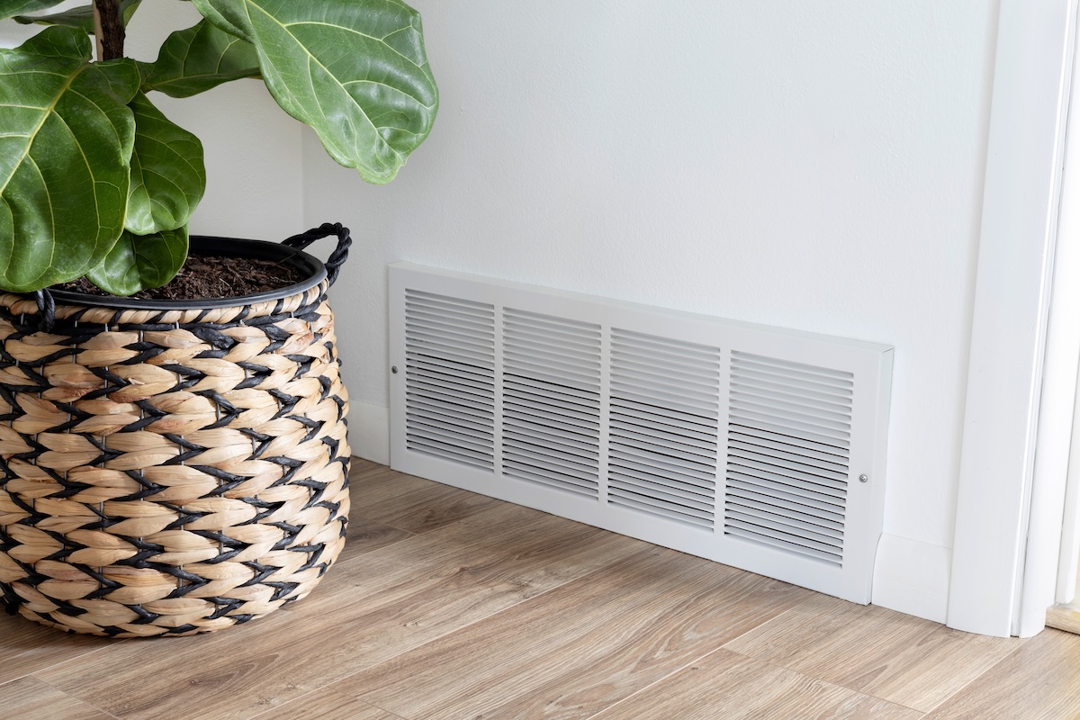 A plant in a seagrass planter is positioned in front of a low air conditioner vent on a wall.