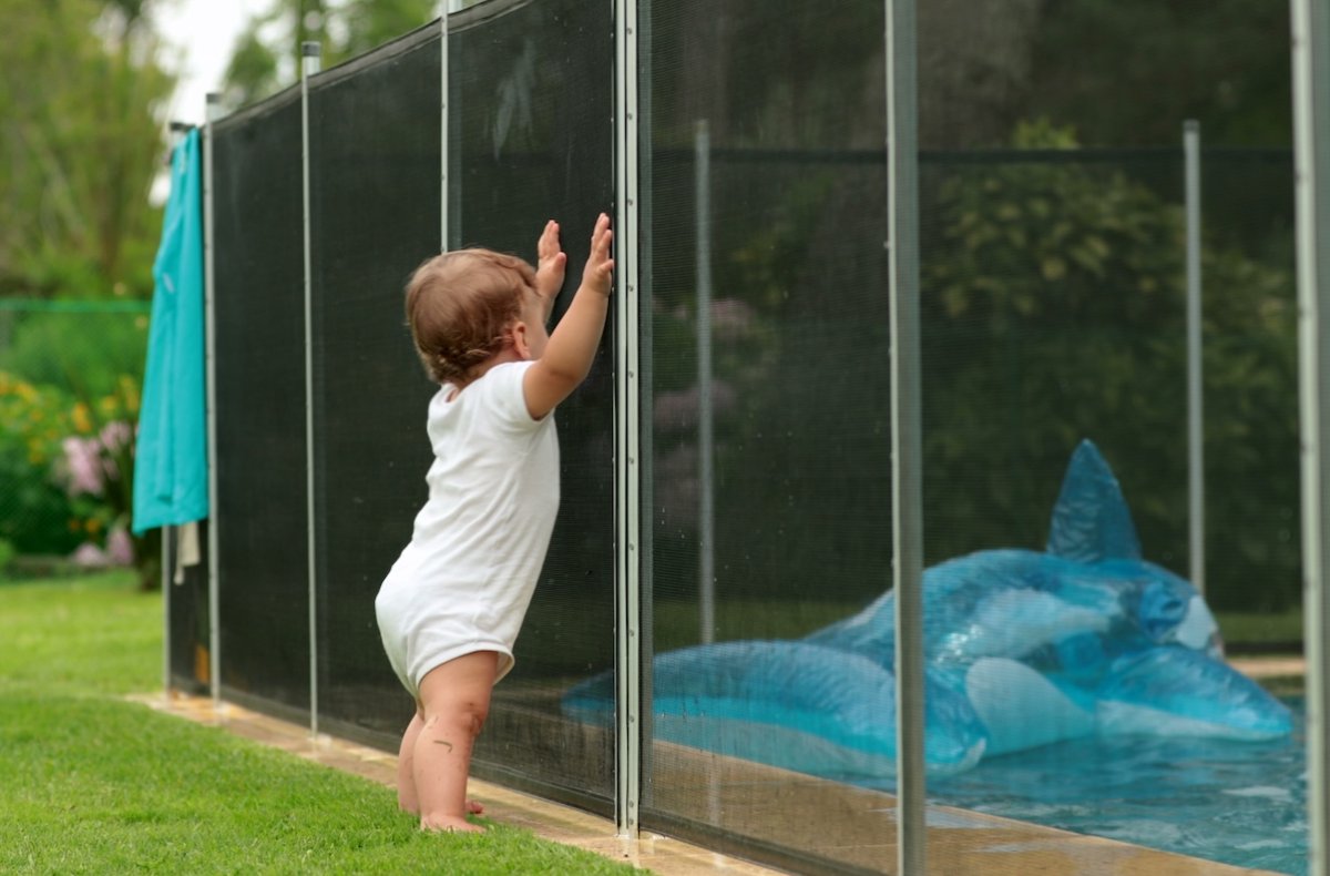 Baby leaning on swimming pool fence  looking in at pool and pool float.