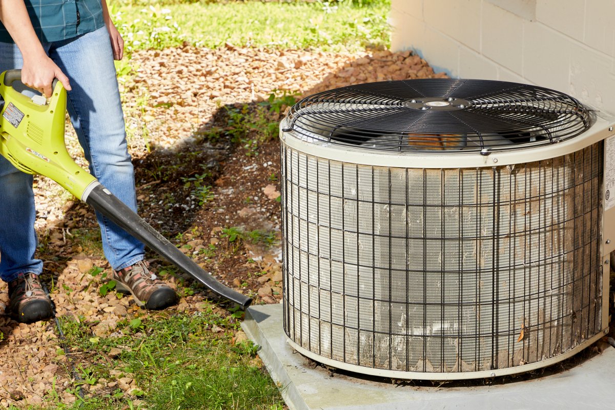 Woman uses a leaf blower to clear debris around air conditioning unit outside.