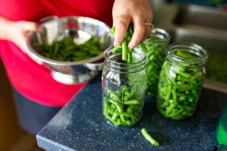 Person wearing a red shirt scoops handfuls of fresh green beans from a colander into glass jars.