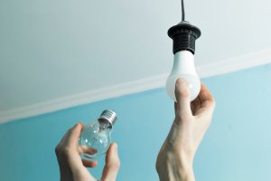 Person holds incandescent light bulb in one hand, replacing it wtih an LED light bulb in an overhead socket.