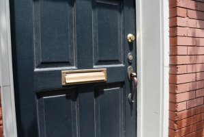 A brass mail slot in a front door painted navy blue, a brick exterior wall adjacent.