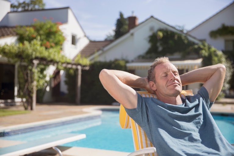 Man stretched out on a lounge chair with his hands behind his head, a swimming pool in the background.