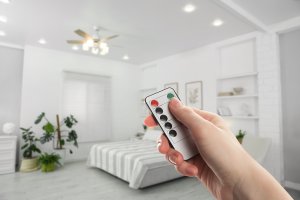 Person points a remote control toward a ceiling fan with a light fixture, in a white bedroom.