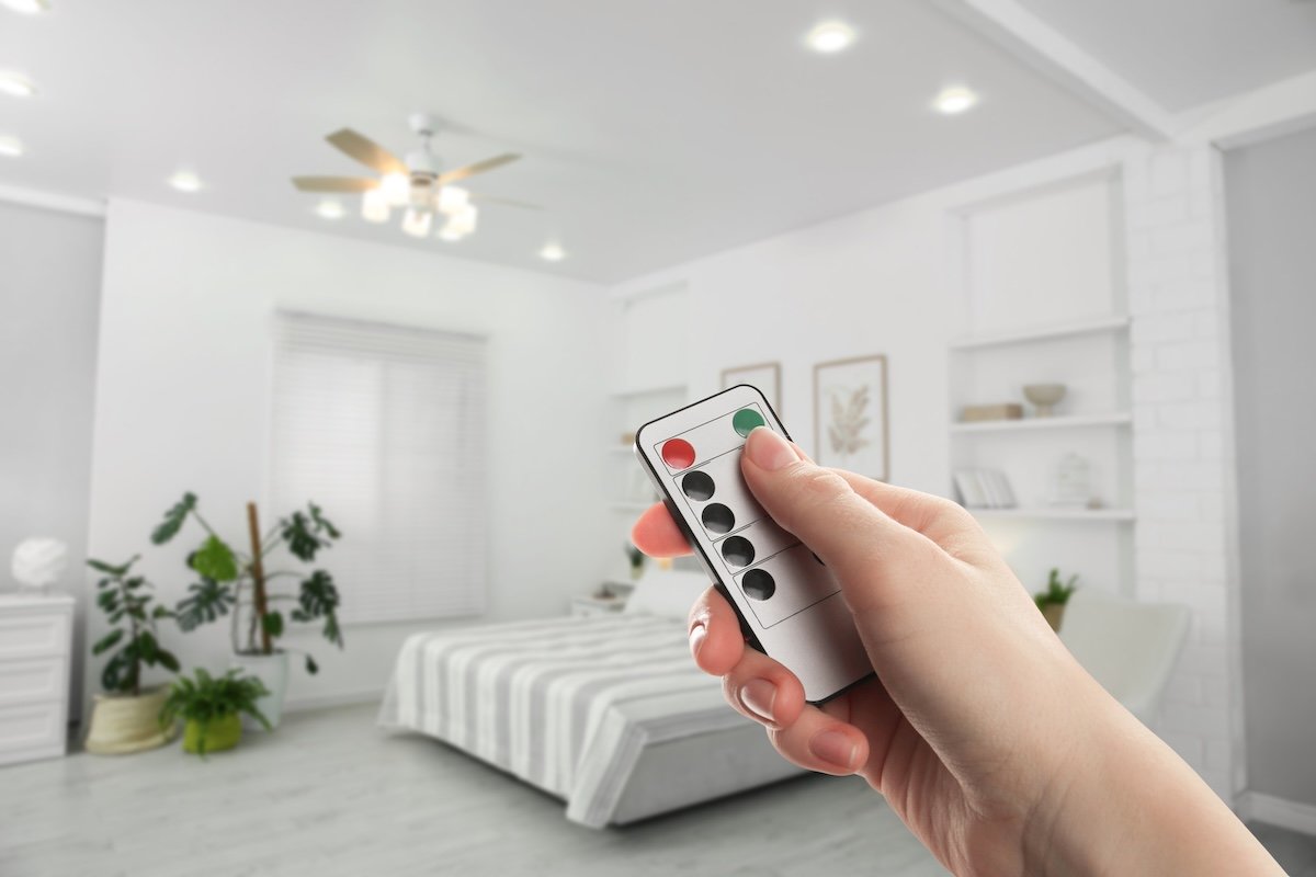 Person points a remote control toward a ceiling fan with a light fixture, in a white bedroom.