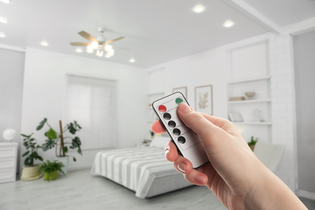 Person points a remote control toward a ceiling fan with a light fixture, in a white bedroom.