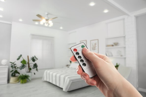 Person points a remote control toward a ceiling fan with a light fixture, in a white bedroom.