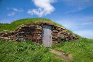 A stone walled root cellar built into a hill with a wood door, a beautiful blue sky above it.
