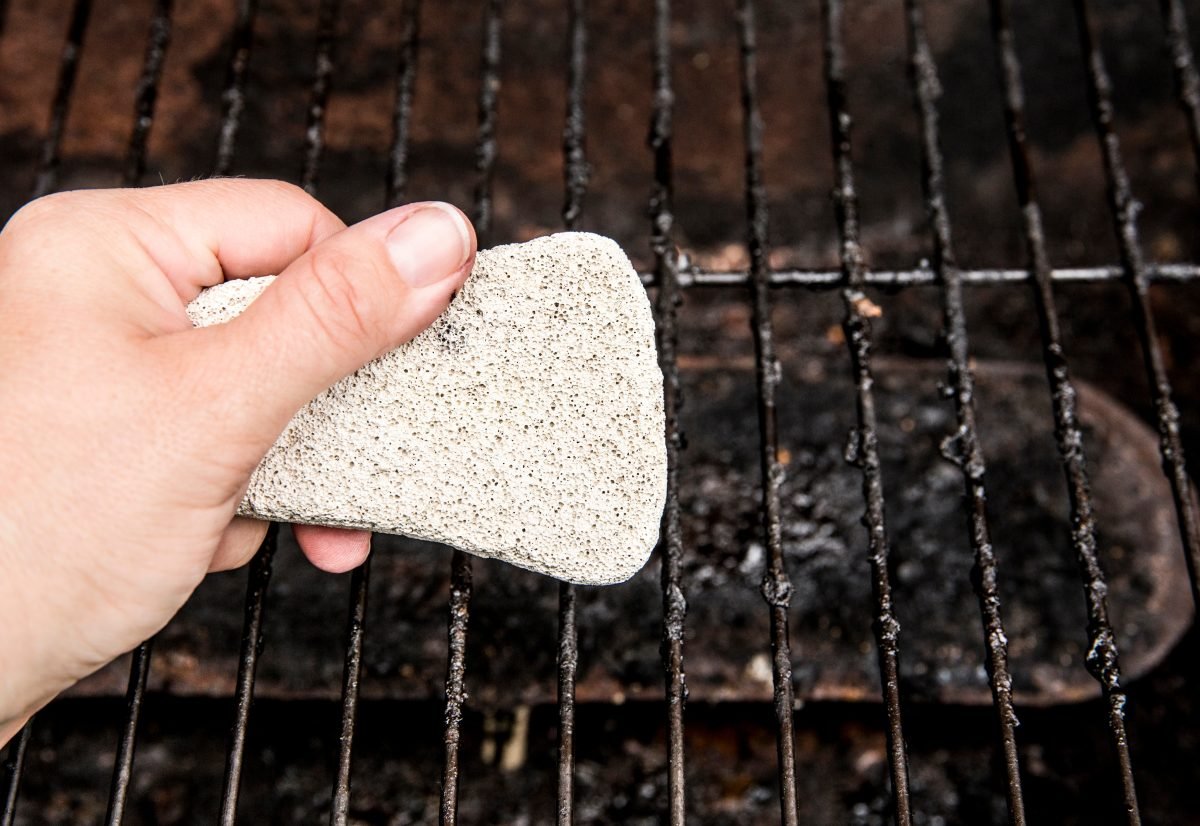Hand using natural pumice stone to scrub charred food deposits and rust from BBQ grill metal grates.