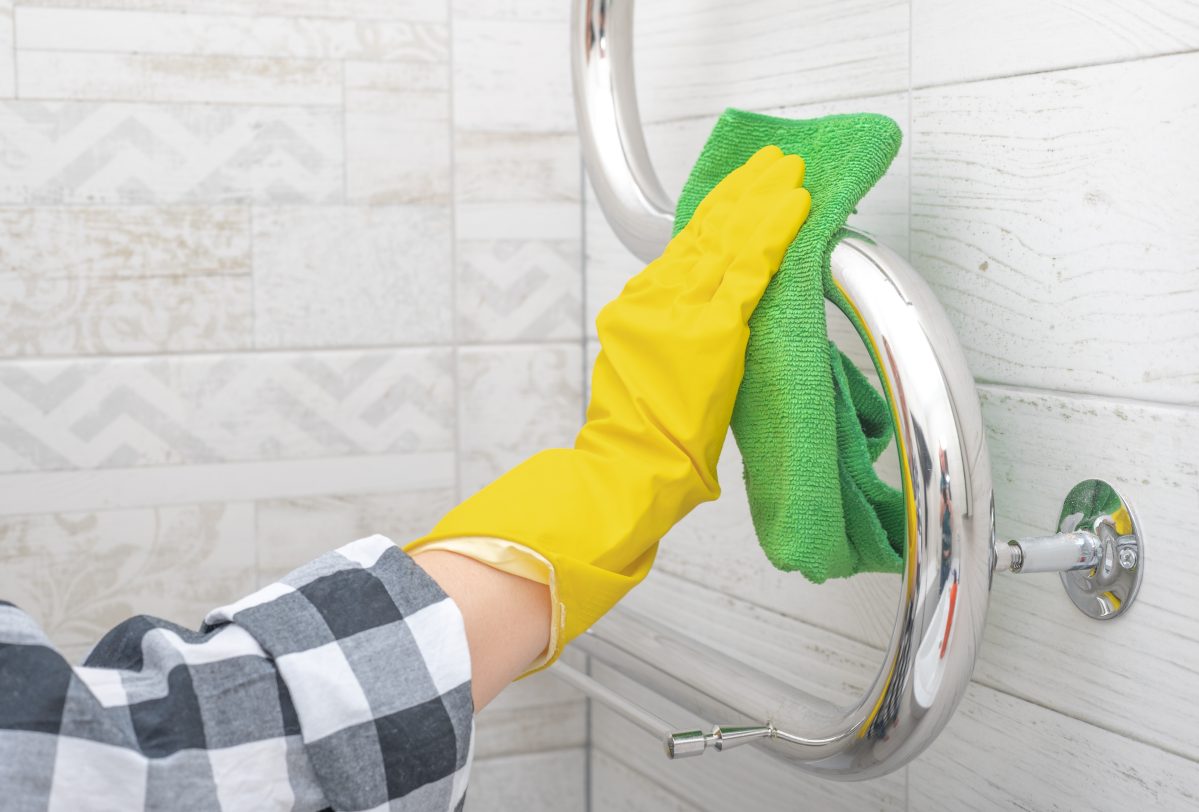 Woman with yellow rubber glove cleaning chrome towel rail in bathroom.