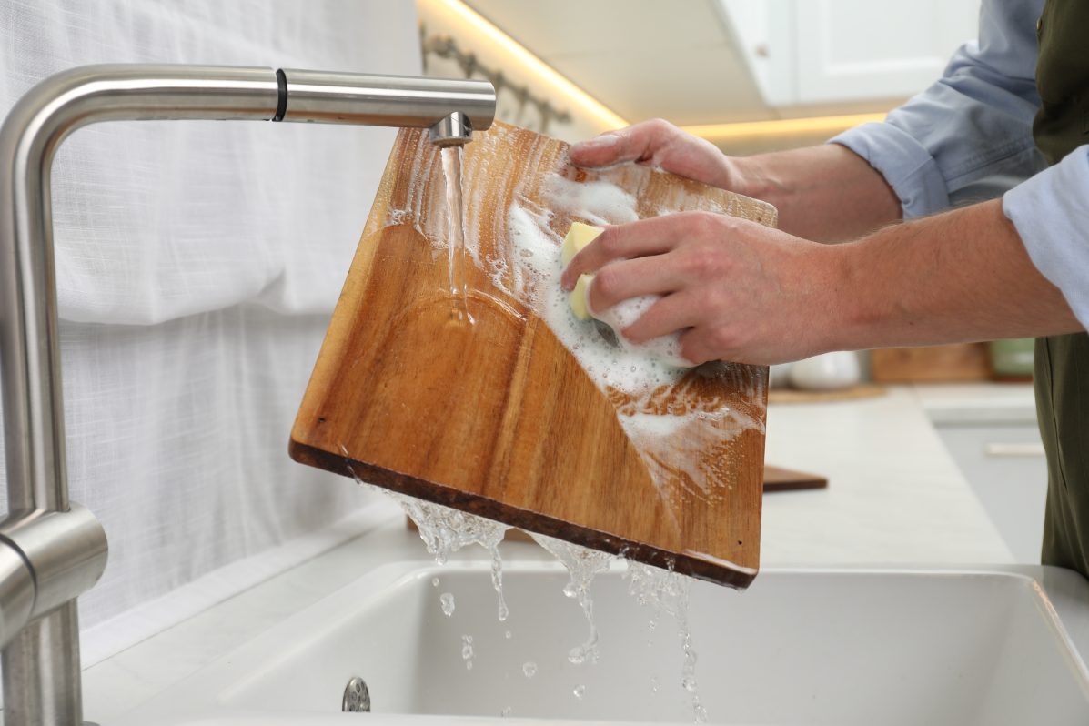 Man washing wooden cutting board in kitchen sink with soap and water.