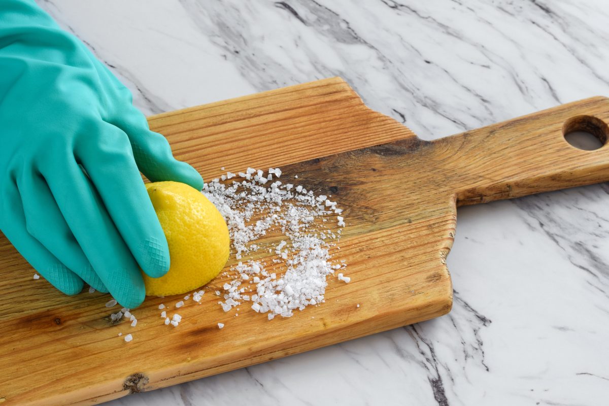 Closeup of person’s hand in protective rubber glove scrubbing wooden cutting board with half lemon and coarse salt.