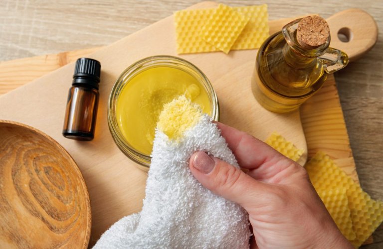 Woman's hands apply homemade beeswax wood treatment polish to restore natural wood cutting board. Beeswax, olive oil and essential oil, soft cloth and mixture in glass jar.
