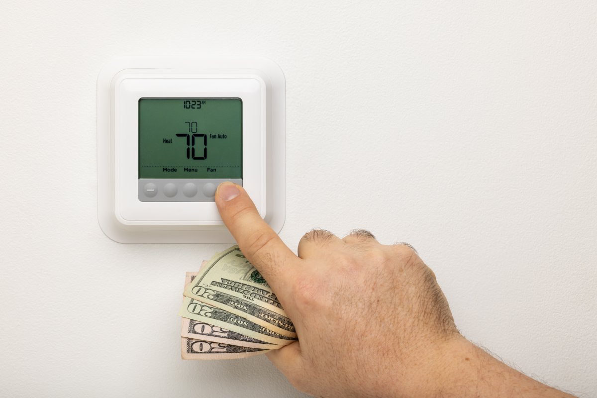 Man adjusting the temperature on a thermostat holding $20s and $50s in his fist