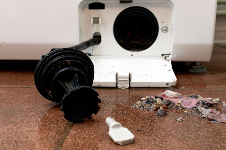 Washing machine drain filter open with debris on floor.