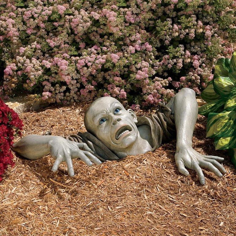 The Design Toscano 8-in Zombie Sculpture climbing out of landscaping mulch in a flower bed